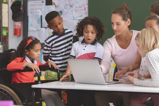 A teacher and group of schoolchildren, including a girl in a wheelchair, gathered around a laptop in a classroom.